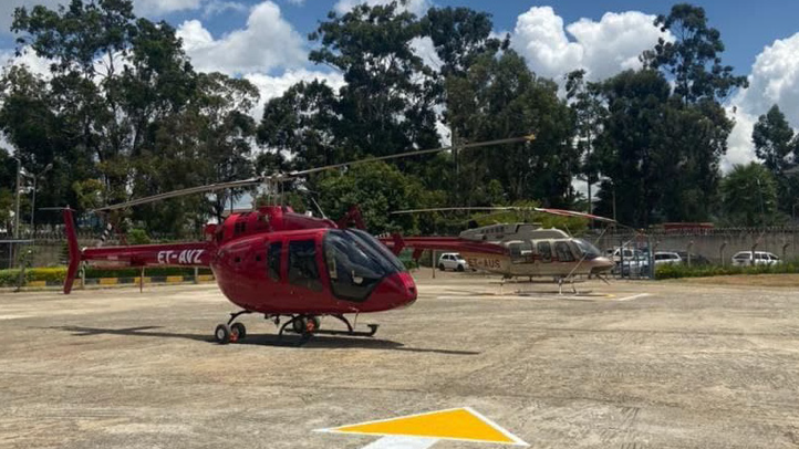 Red helicopter on a helipad with trees and cloudy sky in the background.
